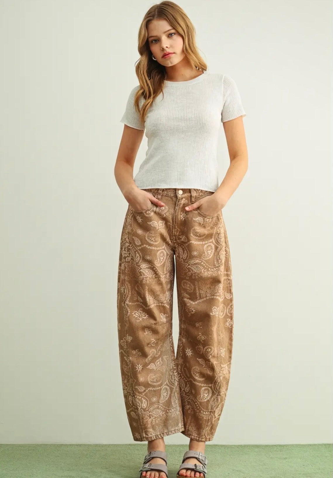 A woman modeling a pair of wide-leg brown pants with a beige paisley pattern, paired with a white short-sleeve top and sandals, standing against a light gray wall.